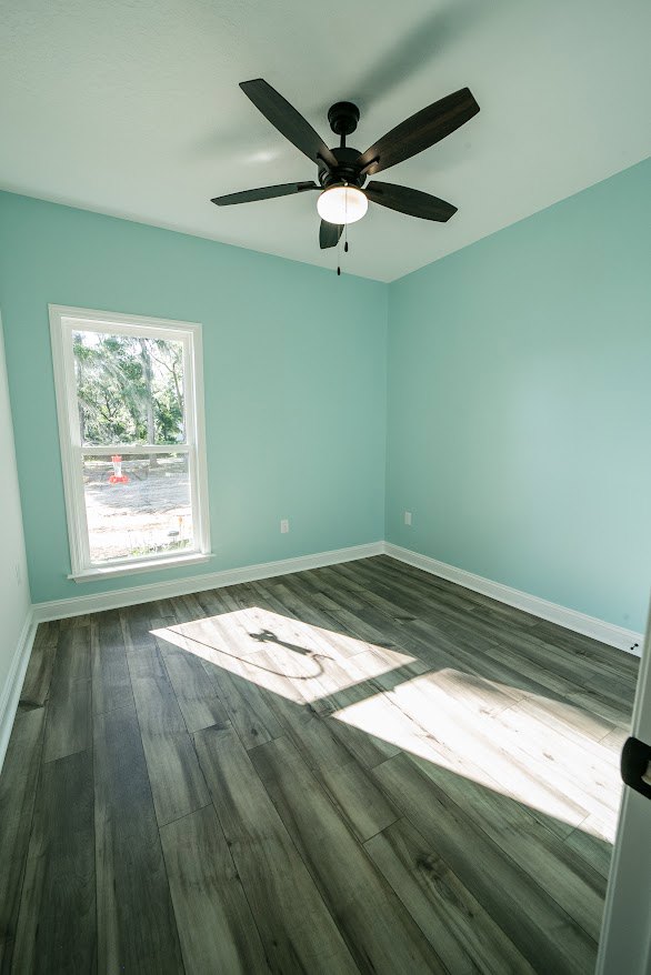 Ceiling fan with light fixture mounted on white plaster ceiling, single window with trim, neutral painted walls, hardwood floor