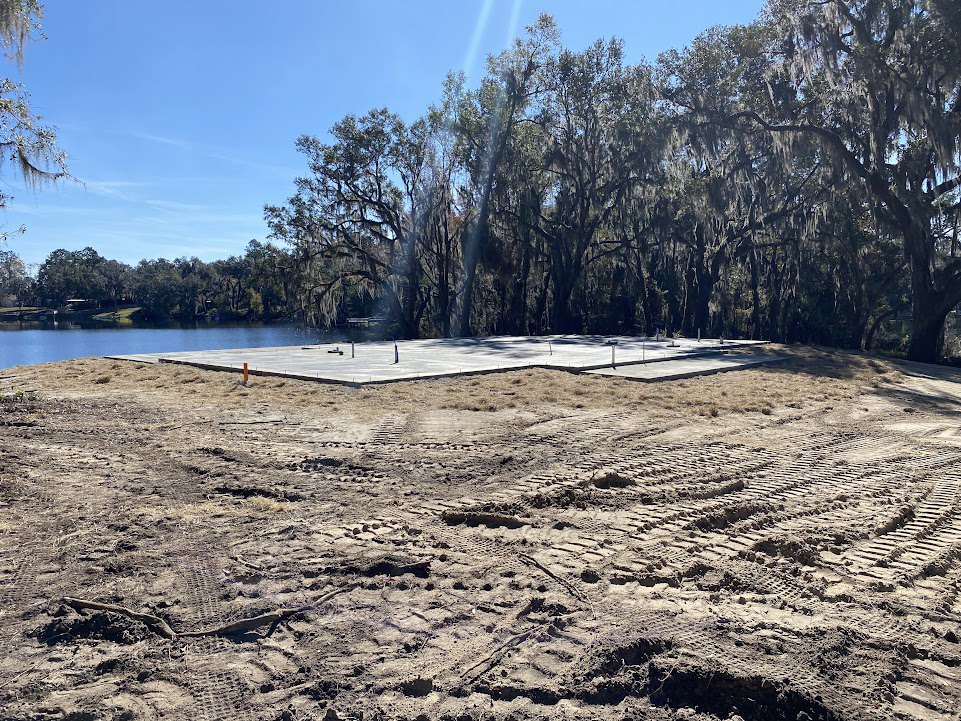 Sandy construction site with tire tracks, snow patches, and grassy ground, bordered by moss-covered trees; lake and dock visible in the background under a blue sky with clouds.