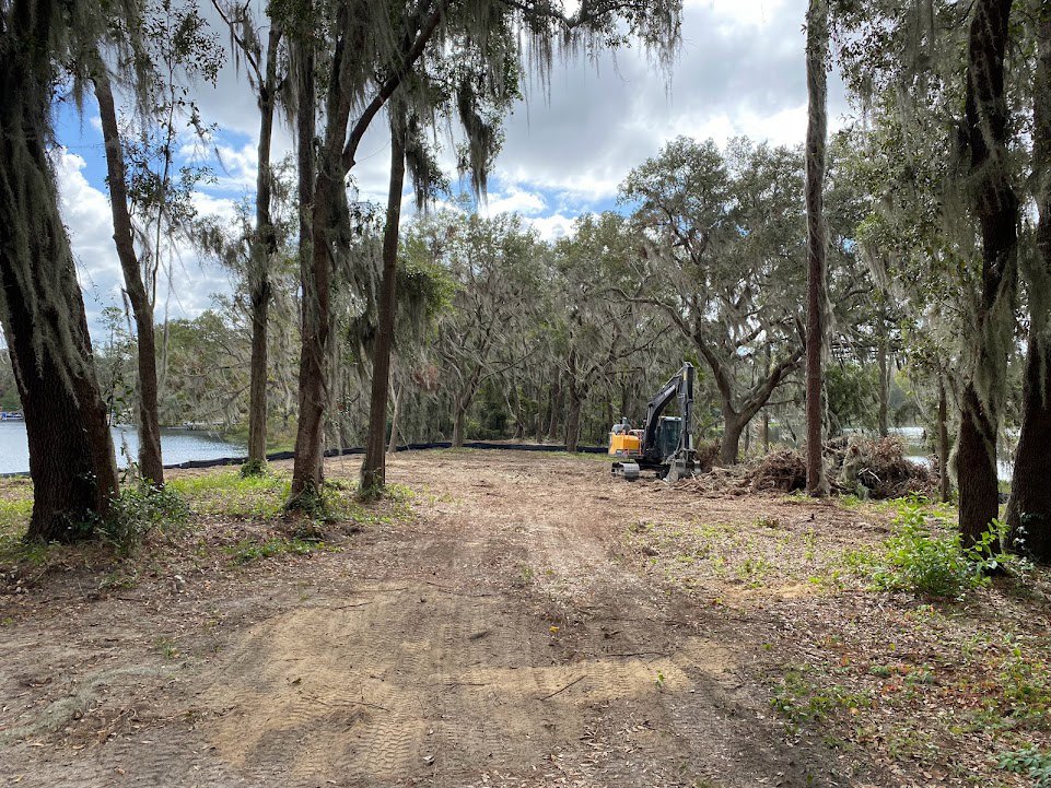 Dirt road with tire tracks winding through dense trees, black tractor parked near yellow construction equipment, grassy ground and cloudy sky visible in the background
