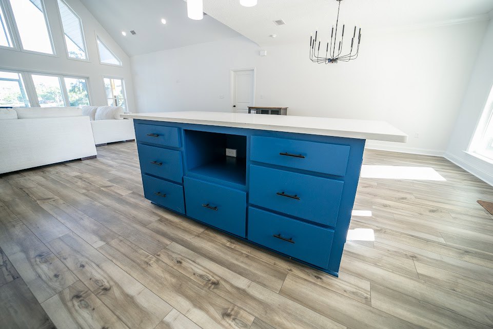 Blue and white kitchen island with black drawer handles, white countertop, nearby blue dresser, candle-style chandelier, white couch, and large windows.