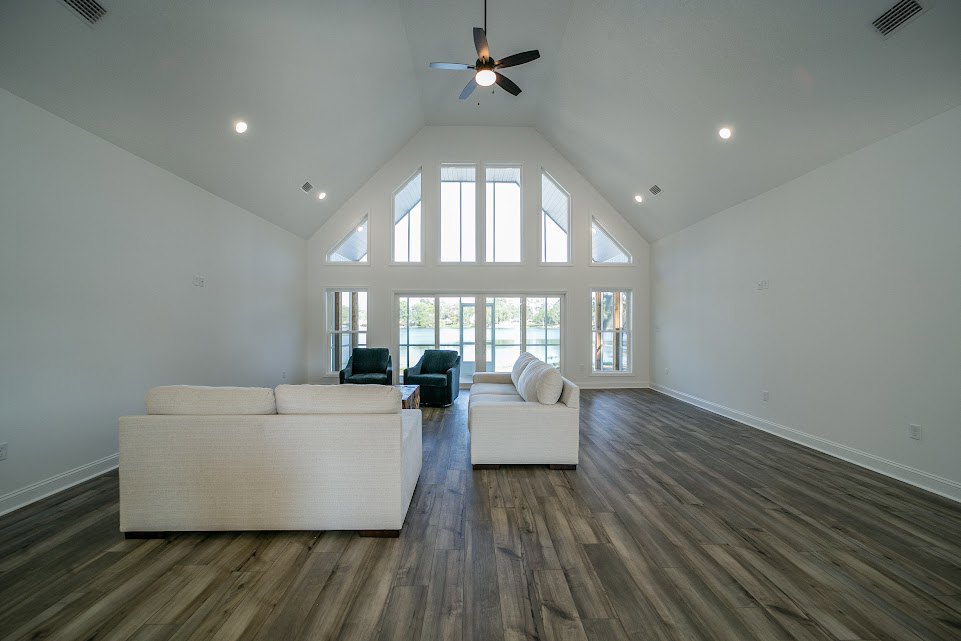 Living room with white upholstered couches, wood laminate flooring, multiple windows, plaster walls, and a ceiling fan with light fixture.