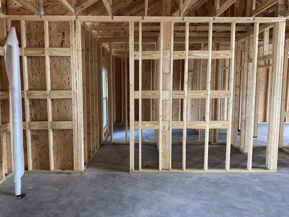 Exposed wood framing and beams over a concrete floor, unfinished interior with visible lumber and wall planks, building insulation partially installed.