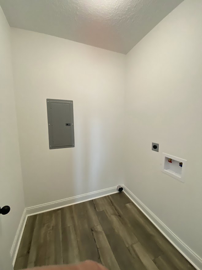 Wood floor with white trim, grey box near wall, grey door with square handle, white rectangular fixture with hole, white plaster walls, ceiling light.