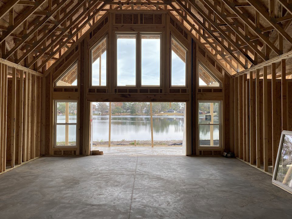 Living room with large white-framed windows overlooking a lake, wood beam ceiling, light hardwood floors, and clear sky visible through glass.