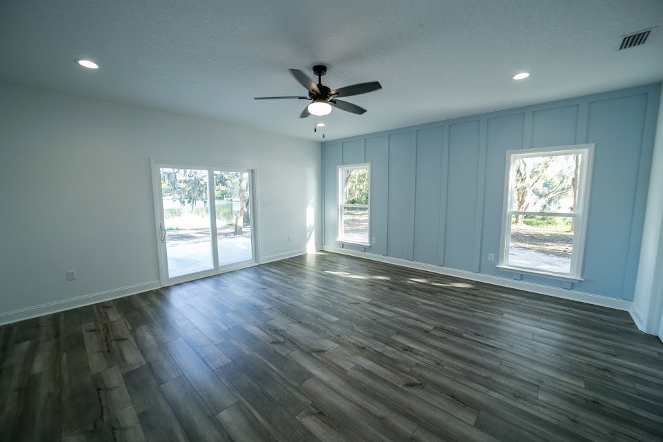 Wood flooring, white walls, ceiling fan with light, glass door overlooking lake, window with white frame and view of trees.