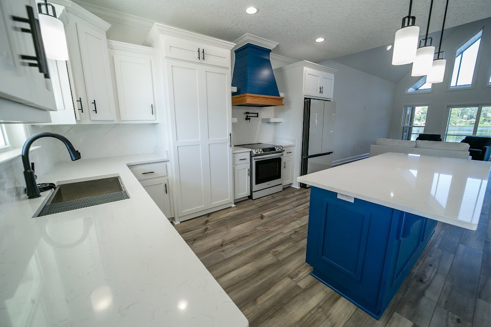White shaker cabinets and blue kitchen island with quartz countertops, stainless steel stove, black curtain, white walls, metal grate on counter, white door with black handles