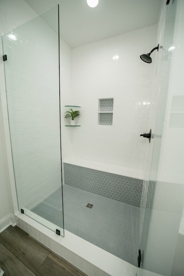 Glass-enclosed shower with black shower head, hexagon-patterned white shelf, and a plant in a white vase on tiled bathroom floor