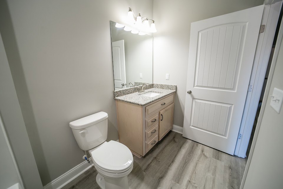 White toilet with closed lid beside white pedestal sink, rectangular wall mirror above sink, silver faucet, light fixture with multiple bulbs, white door with silver knob, light