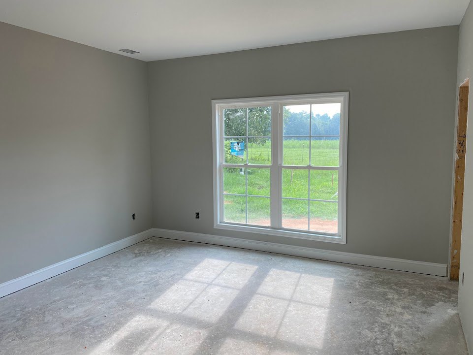 Sunlit room with white plaster walls, black electrical outlet, large window overlooking grassy field and distant trees, light wood flooring