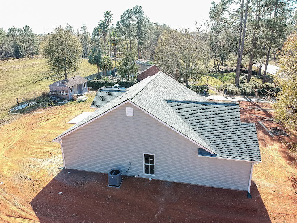 Two-story cottage with gray roof, white siding, and large windows, set beside a dirt road and surrounded by leafy trees under a clear sky