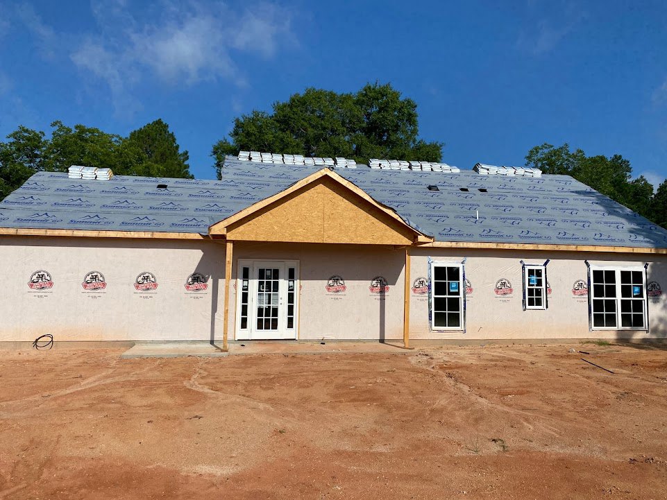 Partially built house with exposed roof, white door with glass panes, window displaying a sign, surrounded by dirt road, wooden post, and mature trees under cloudy sky
