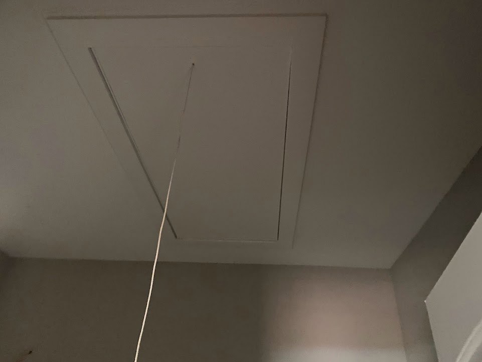 White plaster ceiling and wall with a closed white door, a white cord hanging from the ceiling, and a light fixture visible in the background.