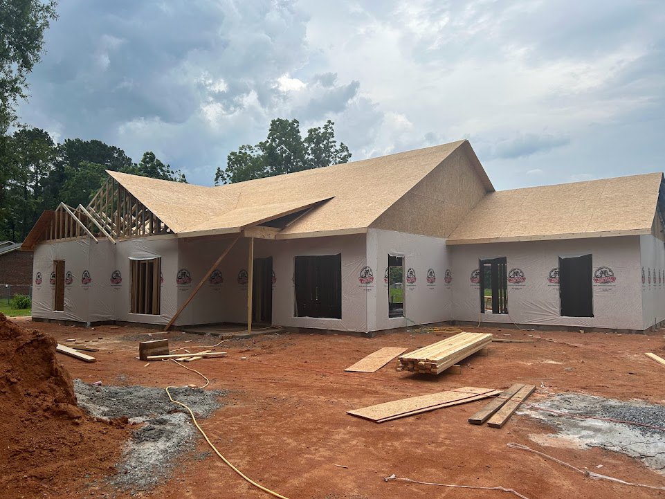 Framed house under construction with exposed wood beams, partially finished roof, black door with silver handles, stack of lumber on pallet, trees and cloudy sky in background