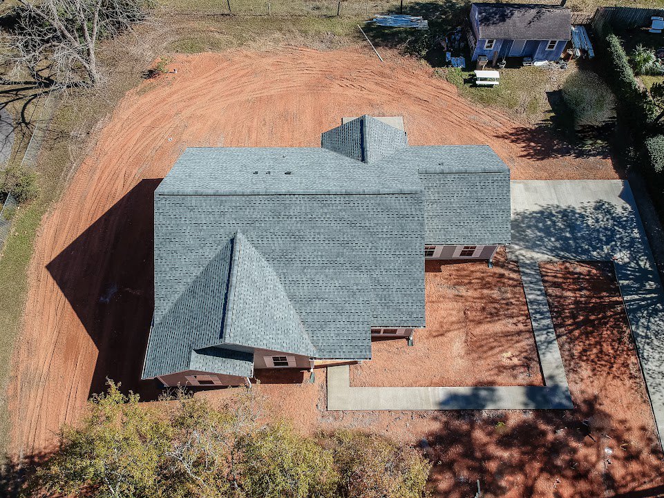 Blue house with shingled roof, concrete driveway, leafless tree casting shadow, grassy lawn, and mature trees surrounding the exterior