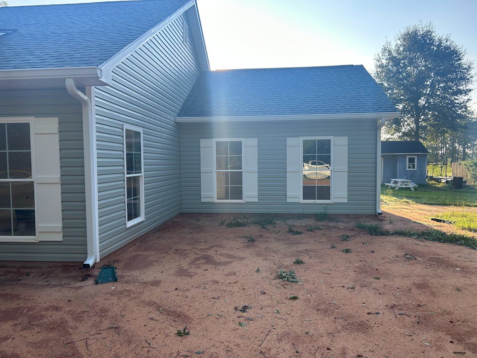 Two-story house with light siding, large windows, front door, landscaped yard with dirt and plants, mature trees in background, parked car visible through window
