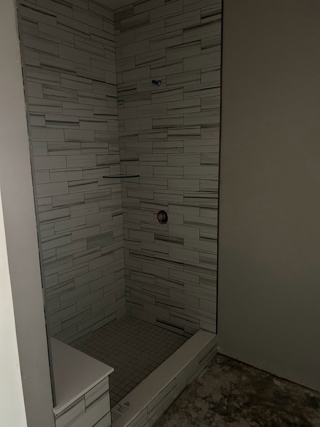 Walk-in shower with patterned tile wall, glass shelf, and adjacent white vanity with drawers; white walls feature black trim, and tile flooring extends throughout bathroom.