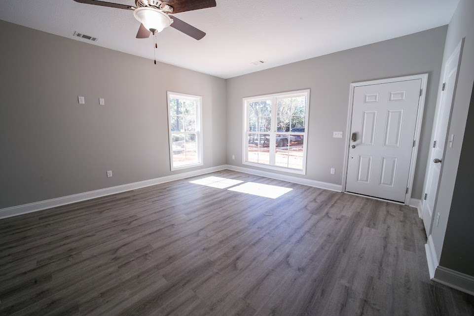 Wood flooring, white walls, ceiling fan with light fixture, window with white frame overlooking parking lot, white door with silver handle