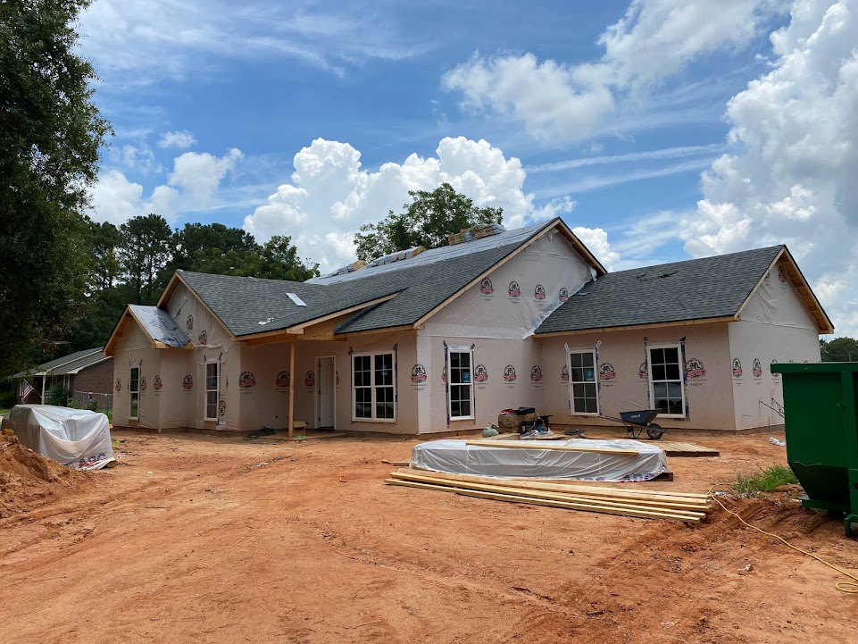Two-story house under construction with white plastic covering, dirt yard, green waste container, pile of wood, and blue sky with scattered clouds
