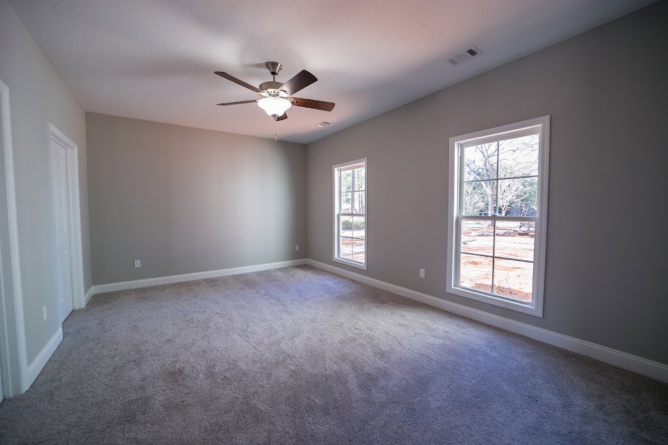 Carpeted room with white walls, ceiling fan with light fixture, large windows framed in white, partial view of trees and construction site outside