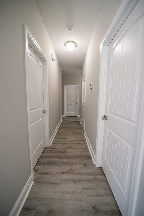 Long hallway with grey walls, white doors featuring silver door knobs, wood flooring, and ceiling light fixture
