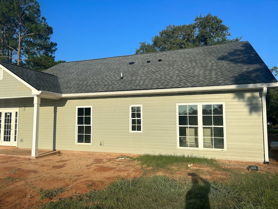 Two-story house with gray siding, white trim, large windows, and a glass-paneled front door, surrounded by a green lawn and mature trees