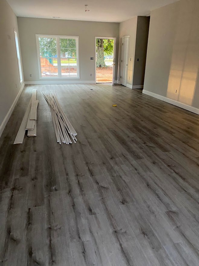 Open room with wide plank wood flooring, large windows framed by white trim, tree visible through glass, white walls, and a white door adjacent to leafy tree trunk.