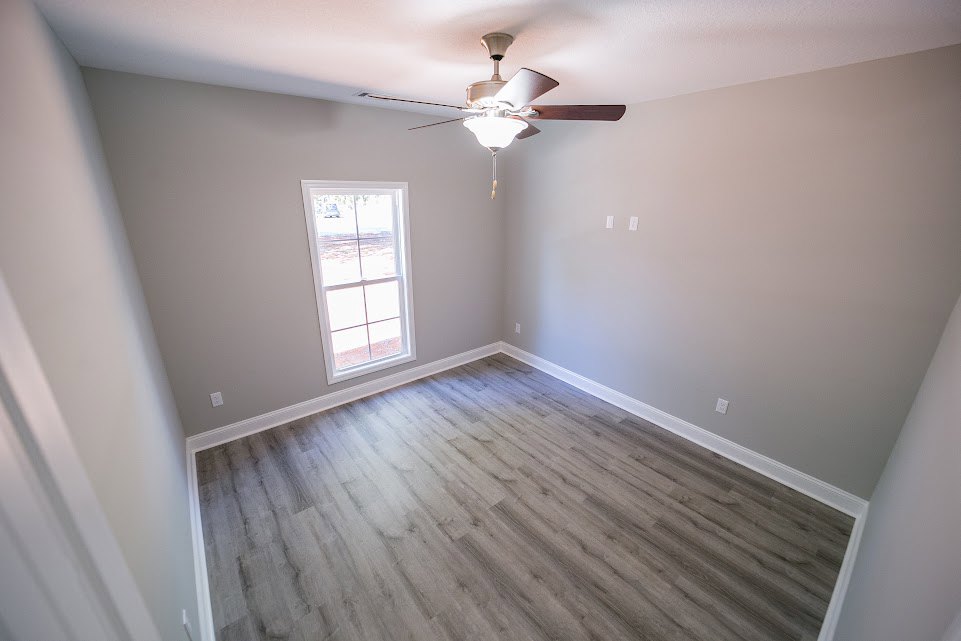 Bright room with white plaster walls, wood flooring, ceiling fan with light, large window framed by molding, sunlight streaming onto floor.