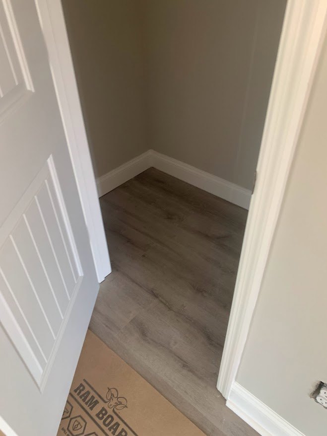 White door with simple molding opens onto a room with light wood flooring and white plaster walls; cardboard moving box sits nearby.