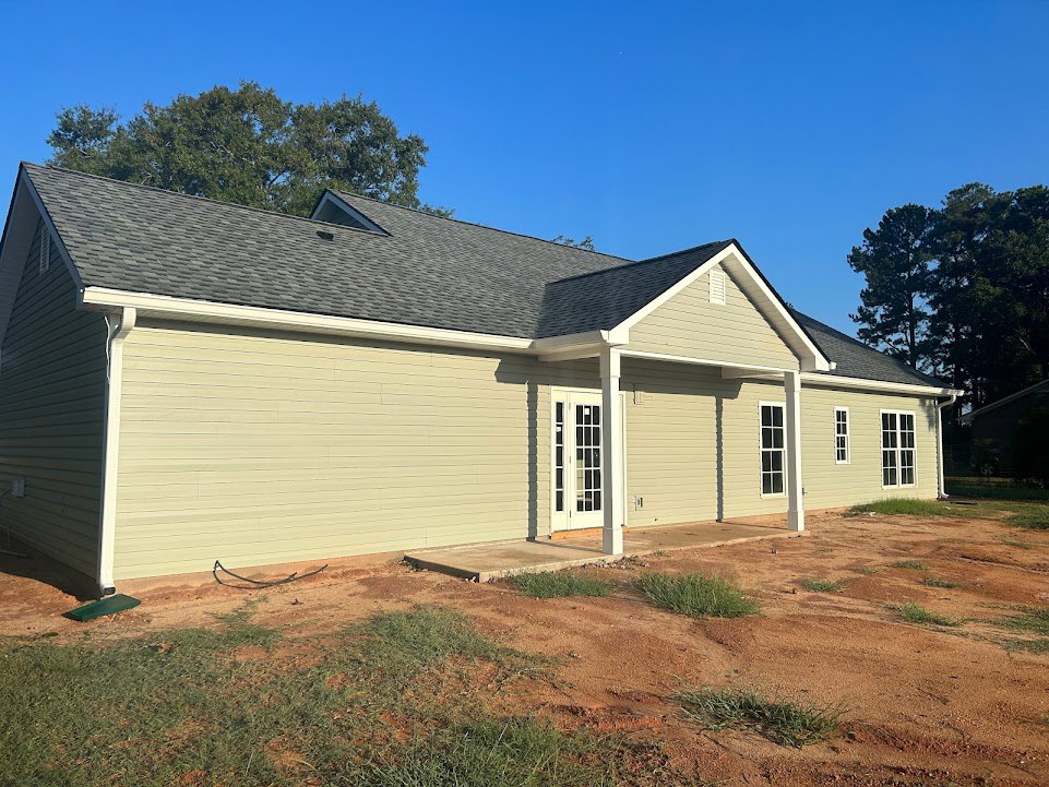 Partially built house with white siding, glass-paneled door, covered porch, dirt patch in front yard, and trees under blue sky