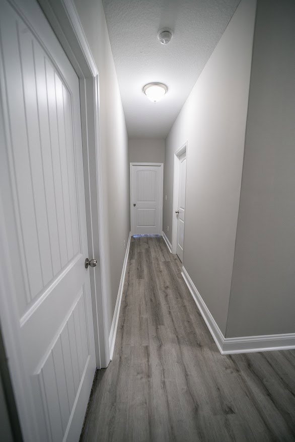 Hallway with smooth white walls, warm wood flooring, white door featuring a black handle, simple base molding, and plaster finishes
