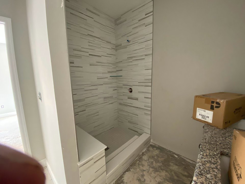 Walk-in shower with glass shelf, plaster walls, wood floor, and a cardboard box with a white label placed near the entrance