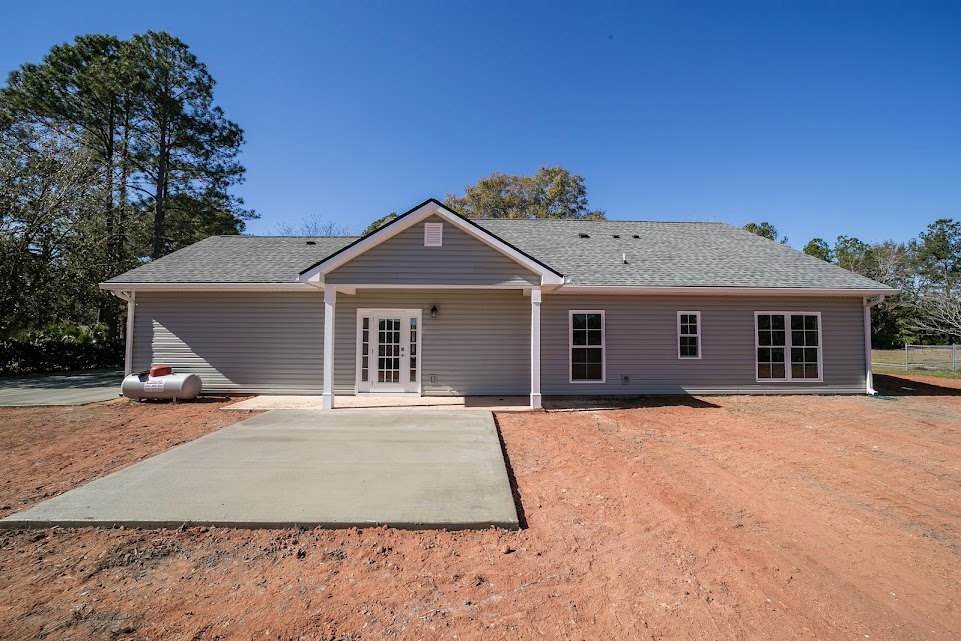 Two-story home with light siding, multi-pane windows, white glass-paneled front door, covered porch, concrete driveway and walkway, mature trees in background