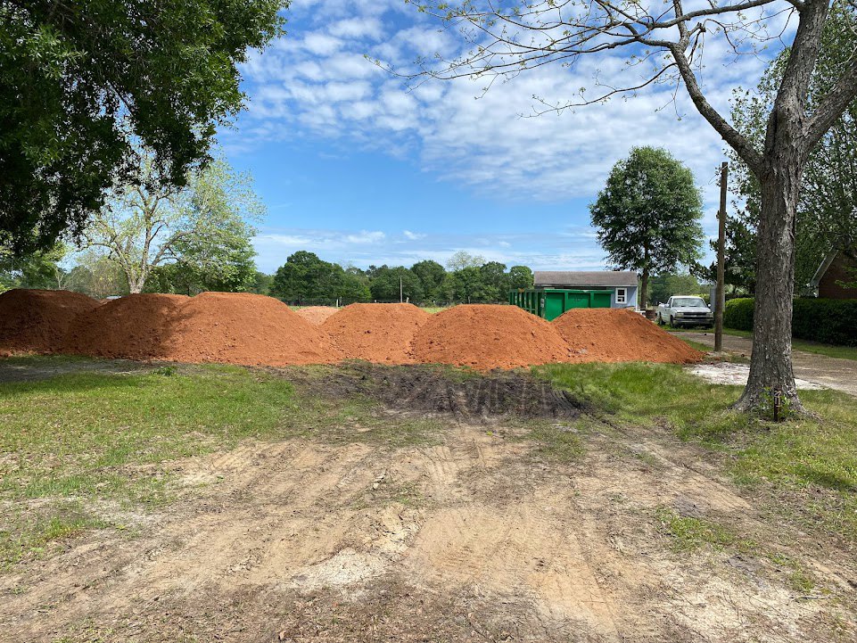 Dirt pile beside a dirt road on a land lot, green dumpster with grey lid nearby, tire tracks across the field, trees in background under blue sky with clouds