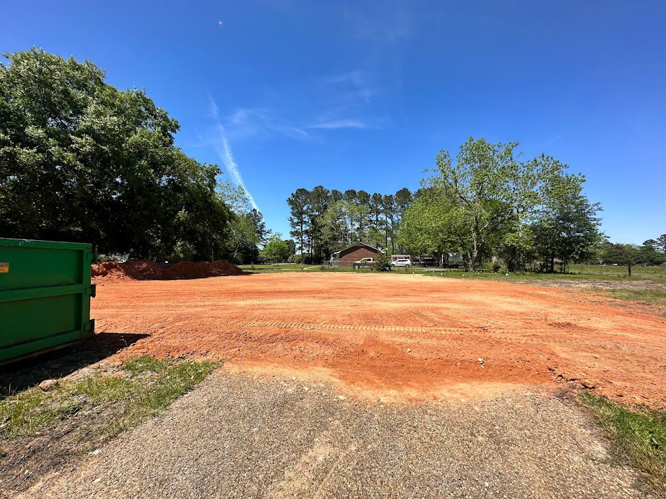 Dirt lot with tire tracks, leafy trees, green waste container with yellow label, building partially visible in background, blue sky with scattered clouds