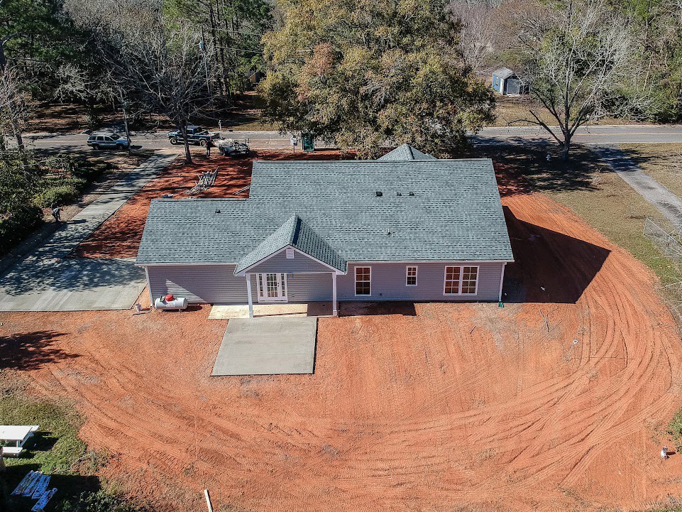 Two-story cottage-style home with gray shingle roof, large windows, concrete driveway, mature leafy trees, and picnic table under white canopy in front yard