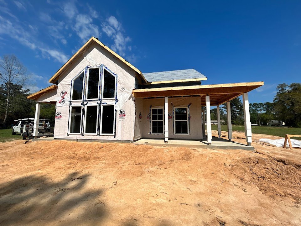 Two-story house under construction with exposed framing, plastic sheeting, dirt yard, leafless tree, and white utility pole in foreground