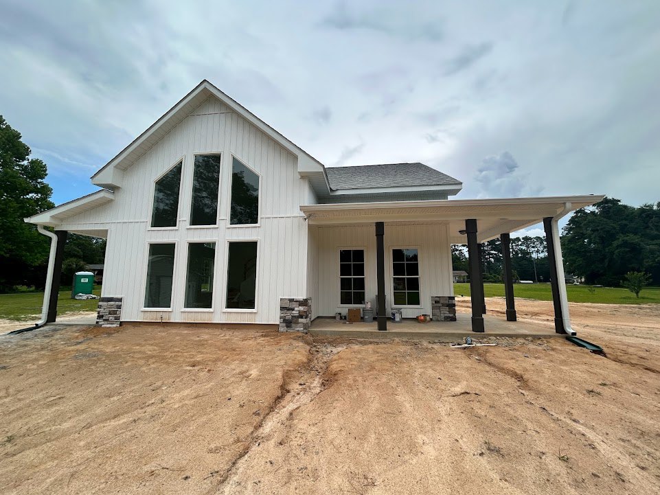 White house under construction with chimney, dirt and grass in foreground, white-framed windows reflecting trees, visible crack in ground near porch.