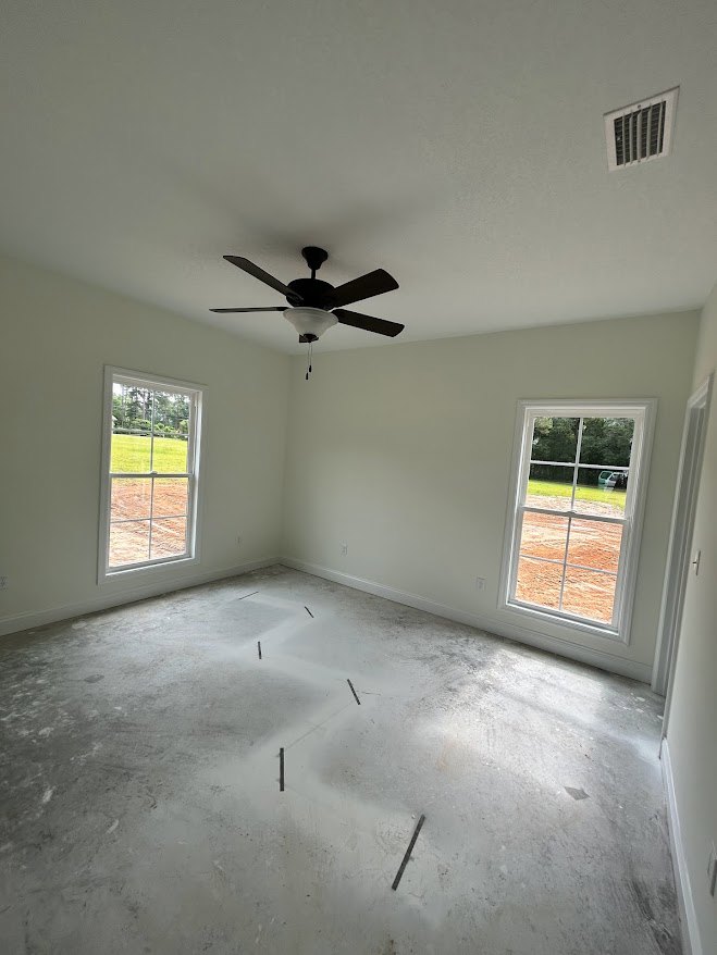 Spacious room with white plaster walls, ceiling fan and light fixture, two large windows overlooking a dirt field, wall vent, unfinished concrete floor with visible holes