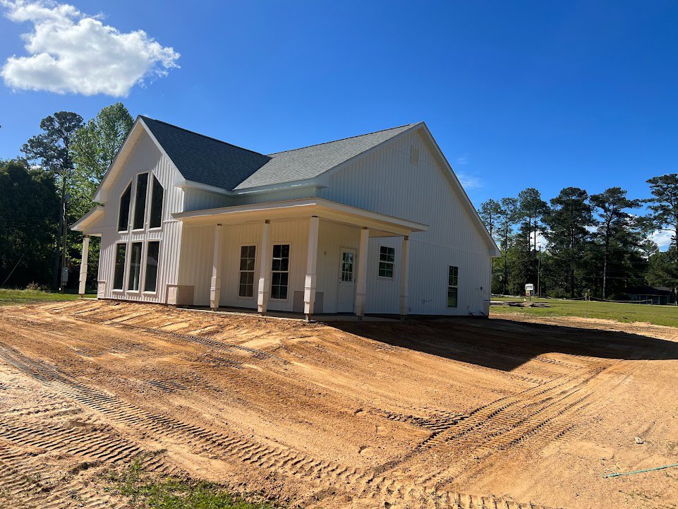 White house with columned entry, dirt driveway with tire tracks, trees and blue sky with scattered clouds