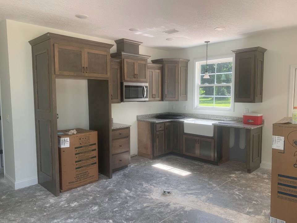 Modern kitchen featuring white cabinetry, stainless steel sink, open microwave door, light streaming through window, brown box with green labels on countertop, and unfinished floor