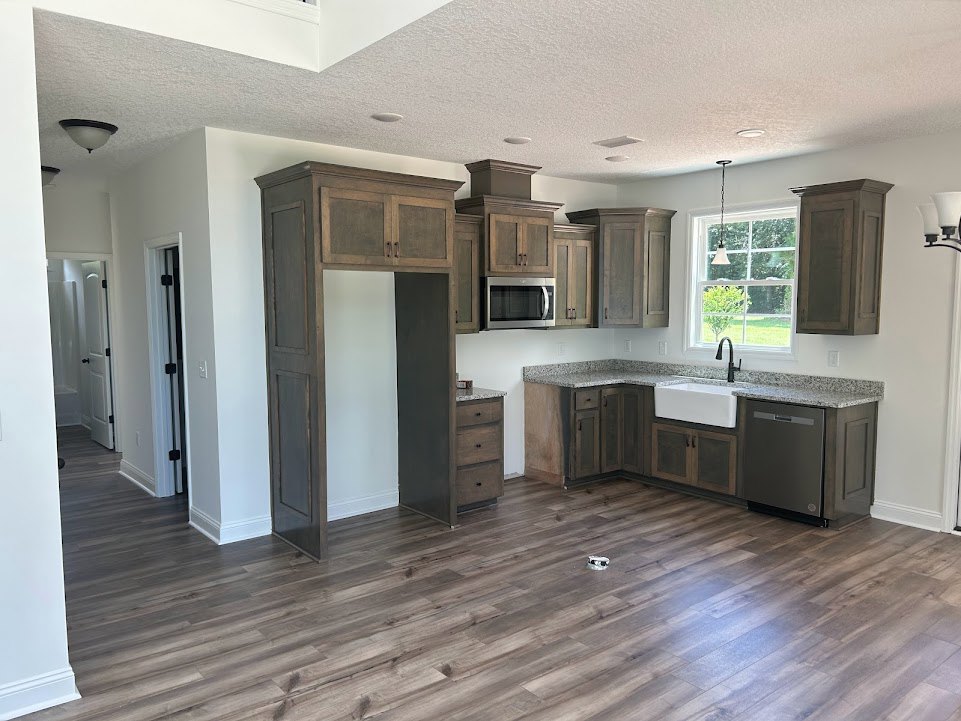 Kitchen with natural wood cabinets, matching wood floor, open microwave door, white rectangular object with patterned edge on floor, window revealing leafy tree outside, door with