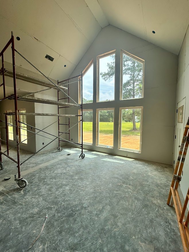 Unfinished room with metal scaffolding, large windows overlooking grassy field and trees, exposed beams, plaster walls, and construction materials on floor