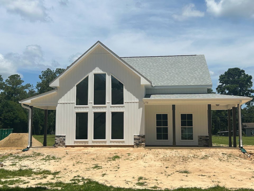 White house with stone roof, white-framed windows, surrounded by dirt field, grass, trees, and green dumpster