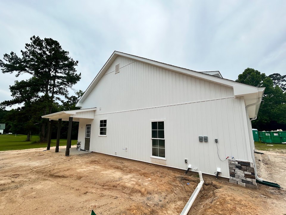 White house with white-framed windows and door, dirt ground in front, green dumpster with white labels, tree beside the building, cloudy sky overhead.