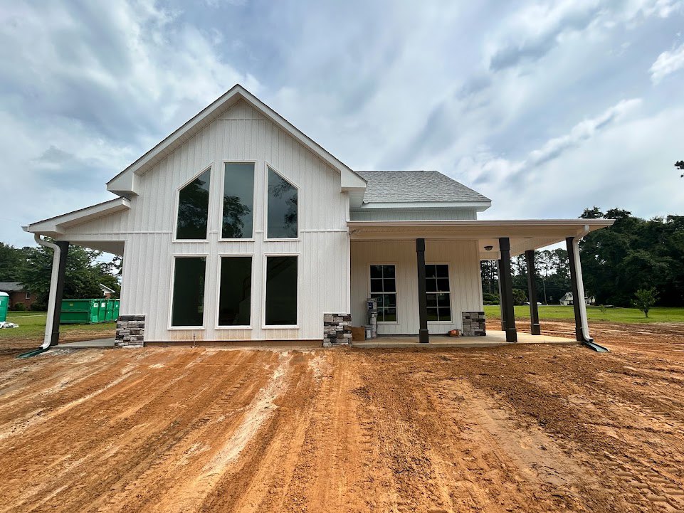 White custom home under construction with multiple windows, surrounded by dirt and mature trees, cloudy sky overhead
