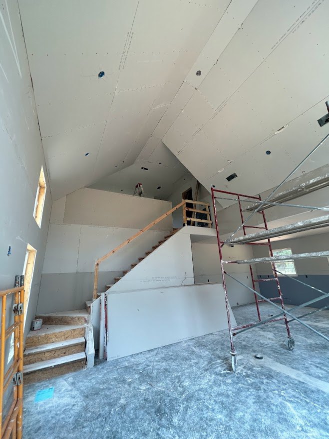 Unfinished wooden staircase with exposed beams, white plaster walls, construction materials, and metal ladder in a partially built interior