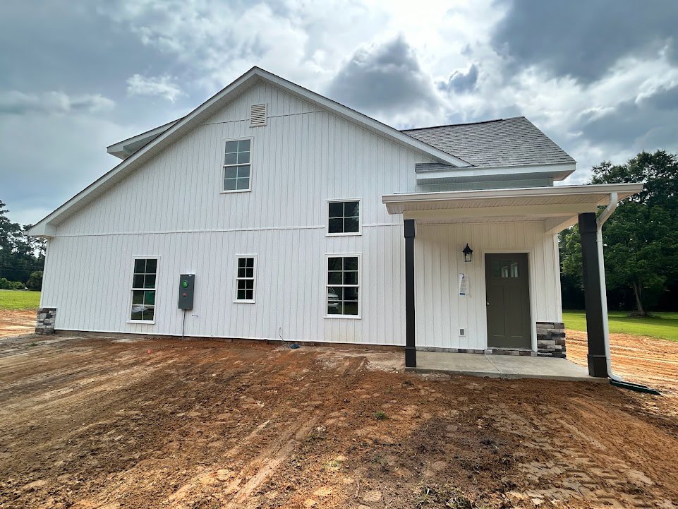 White house with grey roof, brown front door with glass window, white-framed windows, dirt patch in front yard, parked car visible through window.