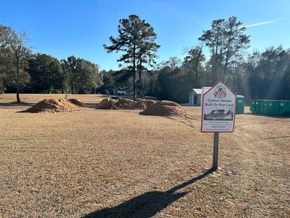Wooden sign mounted on a pole in grassy field with scattered dirt piles, green bins on a table, and a tree with dense branches; shadow of a person's head visible on ground under