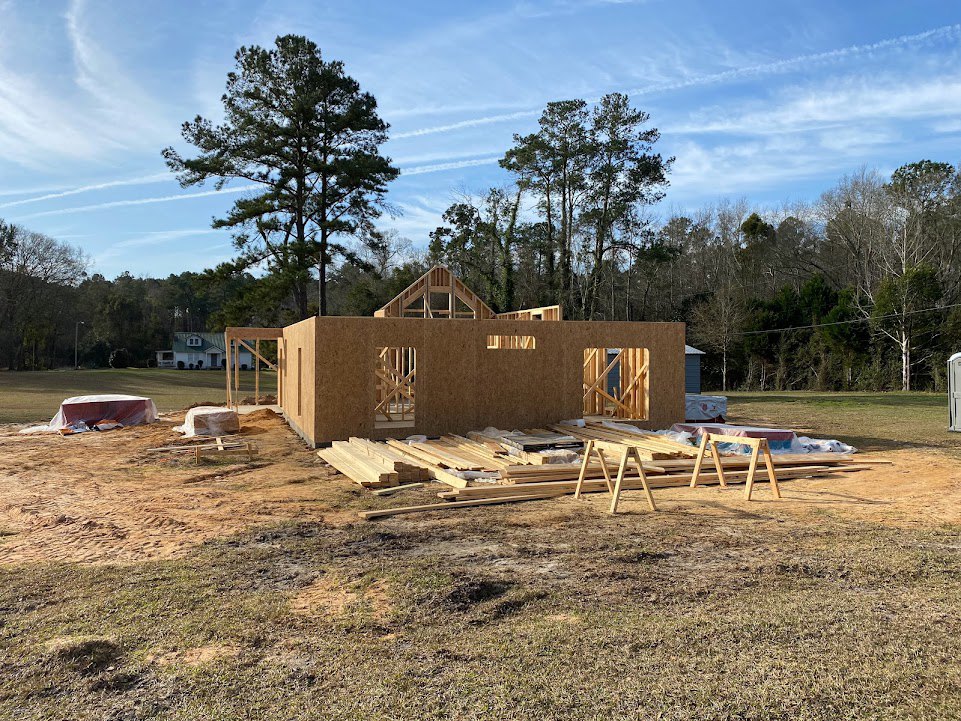 Partially built house with exposed framing and plywood sheathing, surrounded by grassy field, tall trees, scattered lumber, and wooden fence in foreground