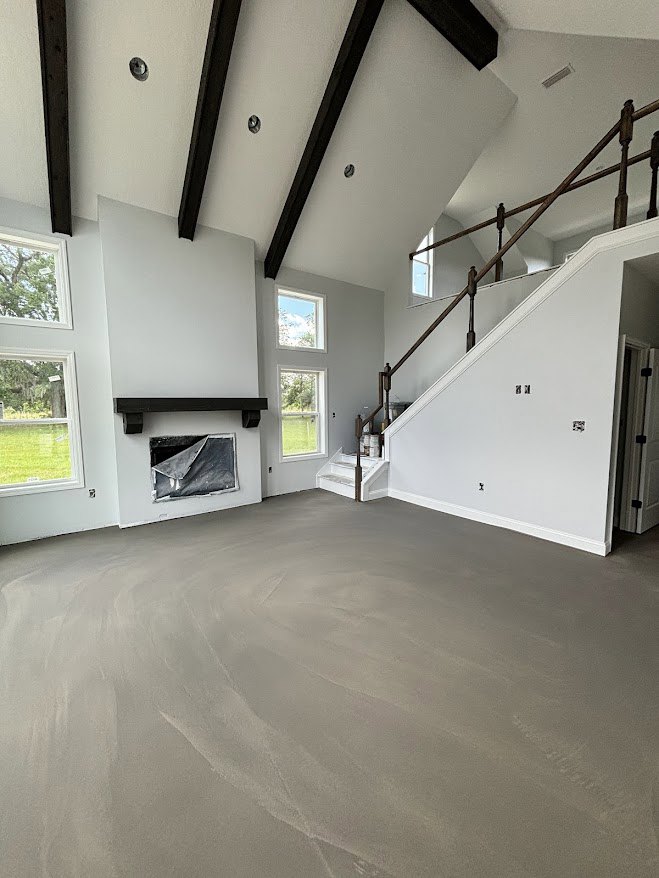 Open living area featuring a modern staircase with metal railing, concrete floor, plaster walls, built-in fireplace, large window with white frame overlooking green lawn, broken